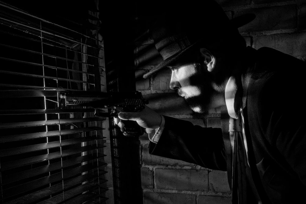 Man aiming rifle through window blinds in dramatic black and white noir style photography.