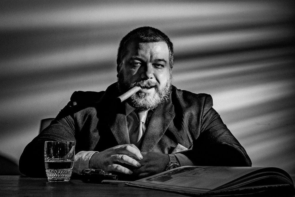 Black and white portrait of a bearded man in a suit smoking a cigar at a table with a glass of water.