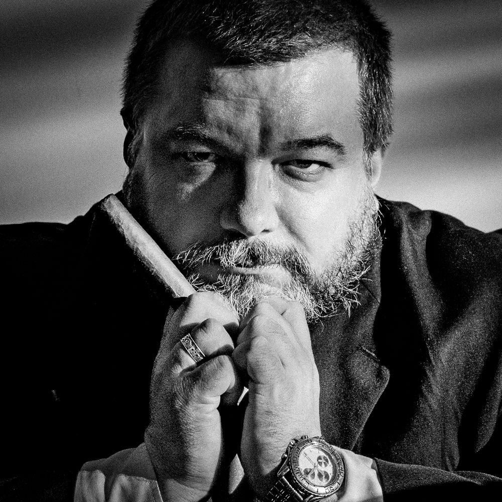 Black and white portrait of a bearded man holding a cigar, wearing a watch, with a thoughtful expression.