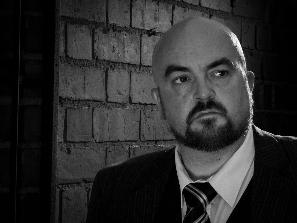 Black and white portrait of a bald man with a goatee wearing a suit and tie against a brick wall.