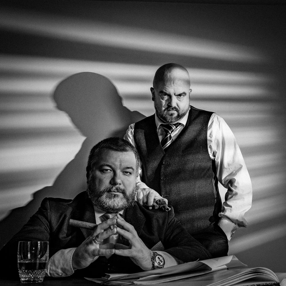 Black and white portrait of two serious men in suits, one holding a cigar, dramatic shadows in background.