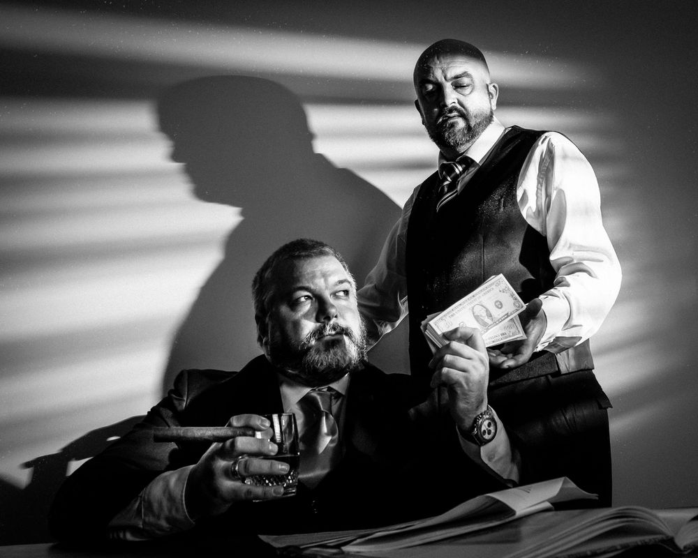 Black and white photo of two men in suits, one holding cash and cigar, dramatic shadows in background.