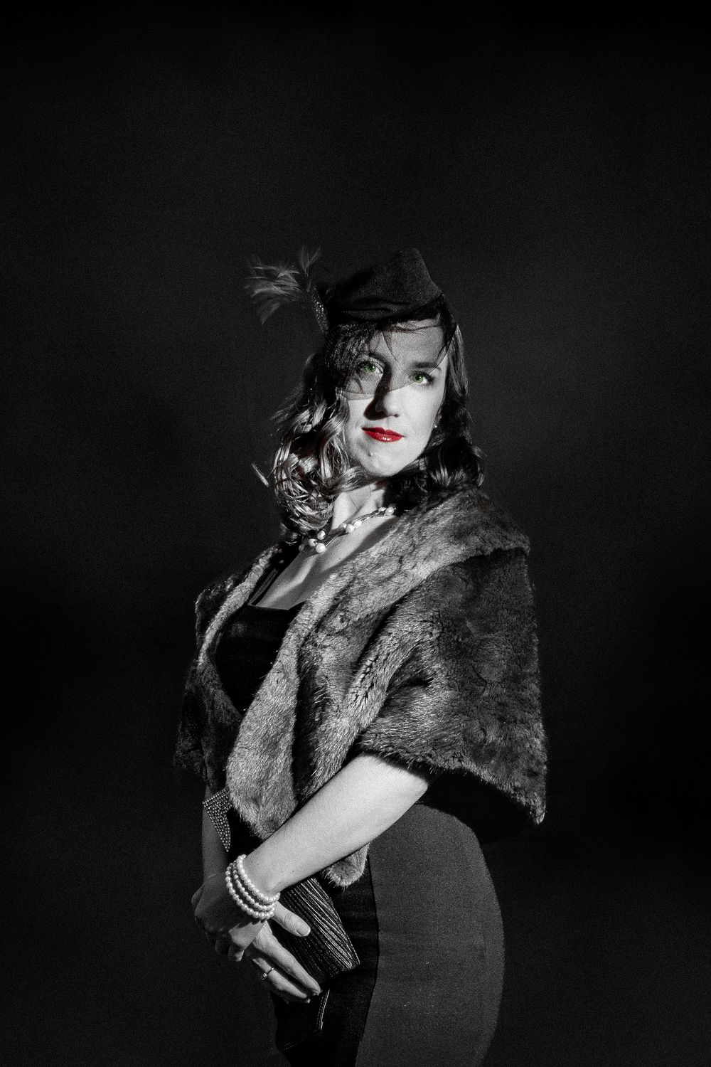 Black and white portrait of elegant woman in vintage fur stole, fascinator hat, pearl jewelry, and red lips.