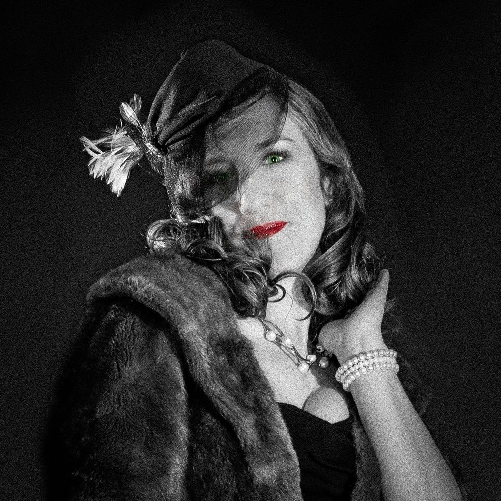 Vintage-style black and white portrait of a woman with green eyes, red lips, pearl bracelet, fur stole, and feathered hat.
