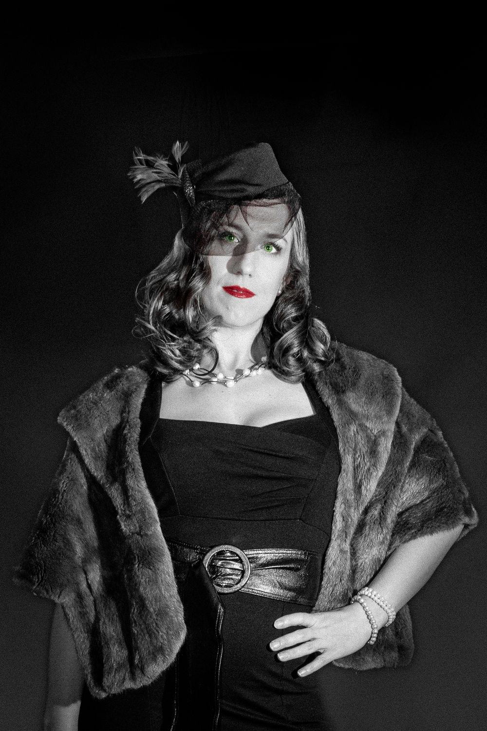 Vintage-style woman in black dress, fur stole, and fascinator hat with red lips and green eyes in black and white photo.