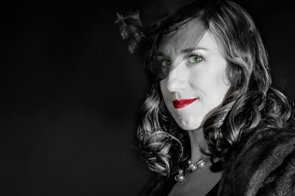 Black and white portrait of a woman with green eyes and red lips wearing a feathered hat and fur coat.