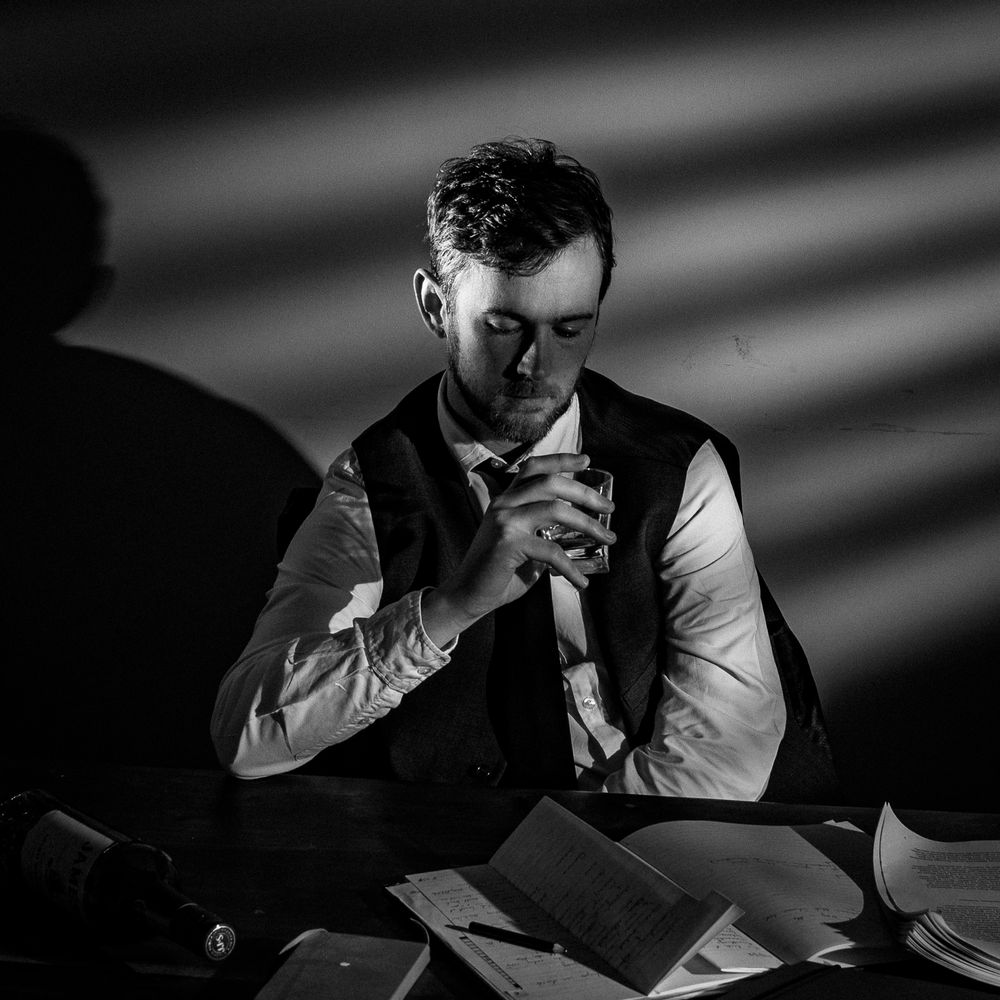 Black and white photo of a young man in a vest sitting at a desk with papers, hands clasped in dramatic lighting.