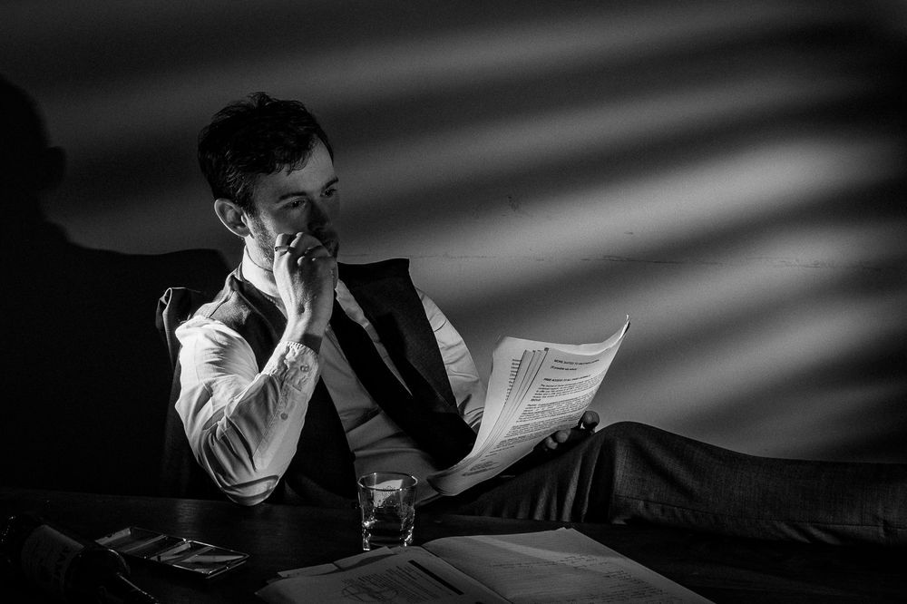 Man in formal vest reads papers while relaxing on a sofa in dramatic black and white lighting.