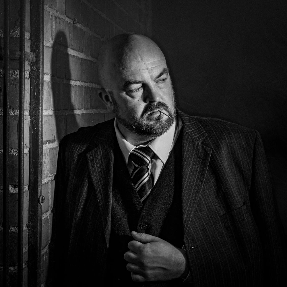 Black and white portrait of a bald man with beard in pinstripe suit leaning against brick wall.