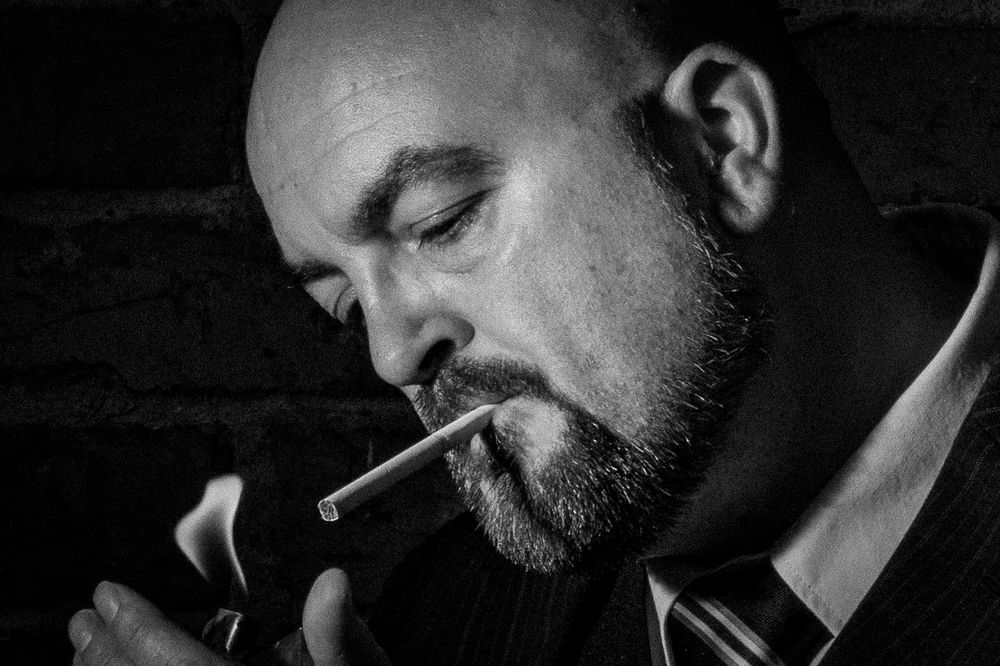 Bald bearded man lighting a cigarette in moody black and white close-up portrait.