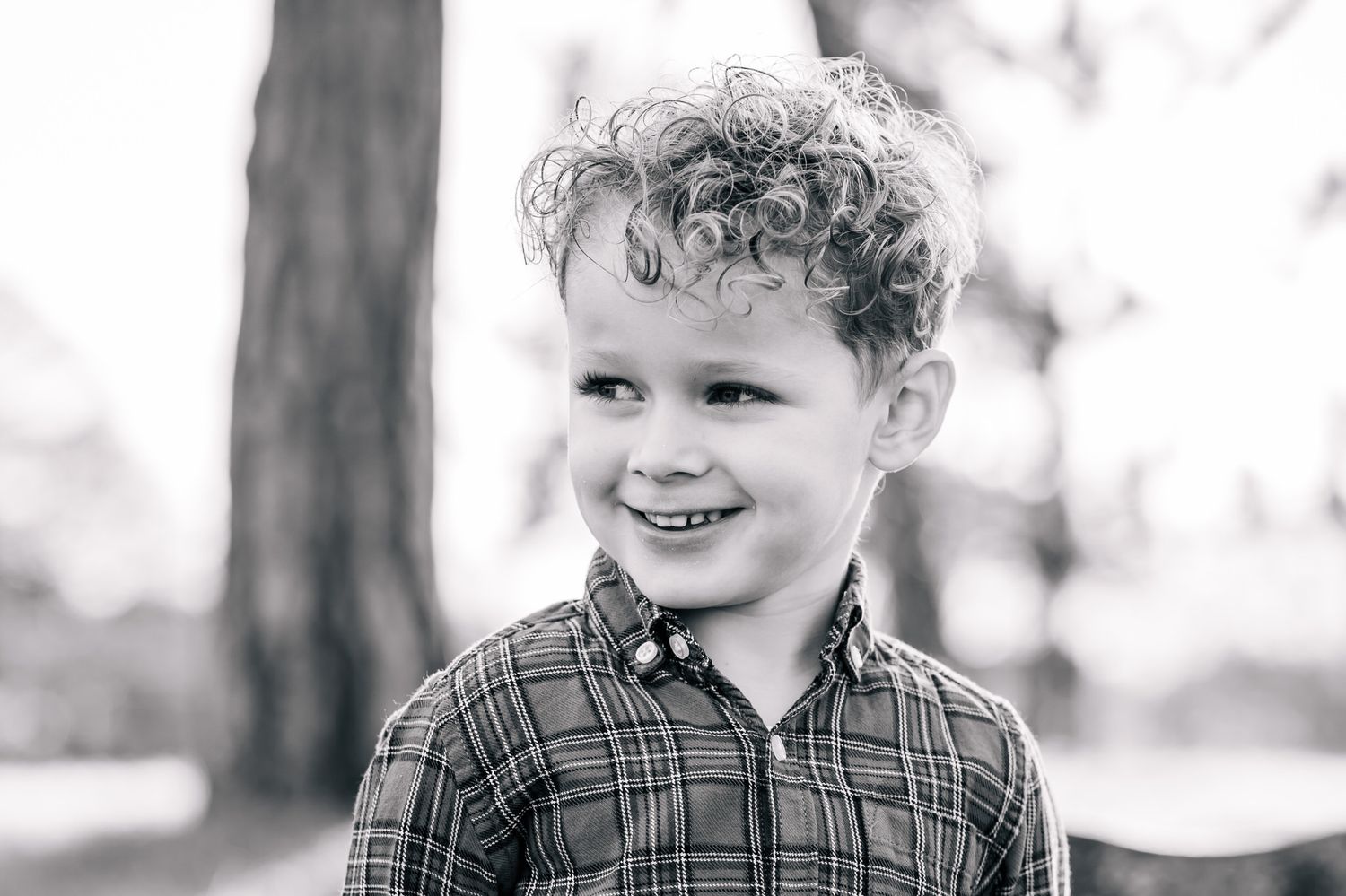 Black and white portrait of a smiling young boy with curly hair wearing a plaid shirt outdoors.