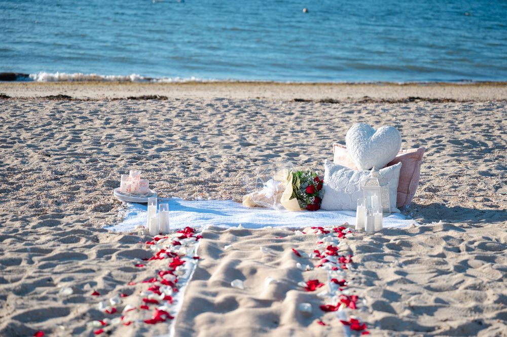 Romantic beach wedding setup with rose petals, candles, white fabric, and a floral bouquet on sandy shore near blue water.