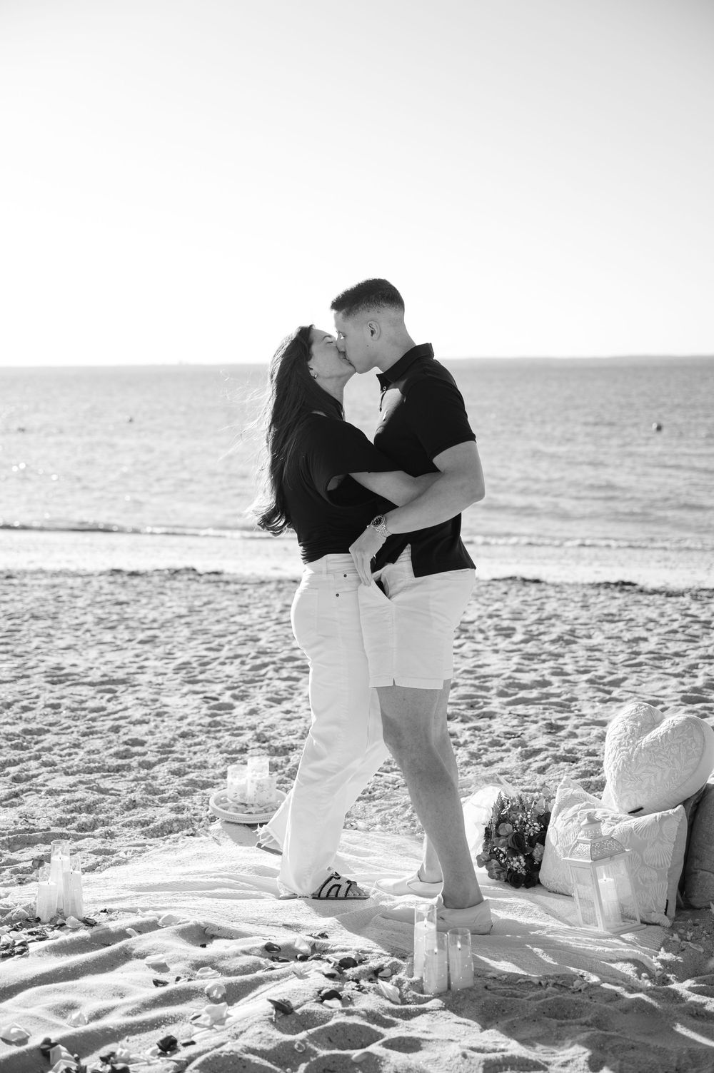 Black and white photo of a couple kissing on a romantic beach setup with candles and flowers at sunset.
