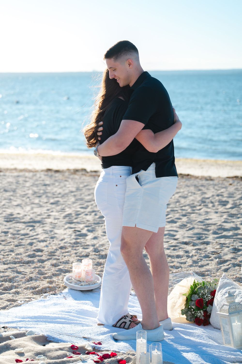 Couple embracing on a romantic beach proposal setup with rose petals, candles, and flowers at sunset.