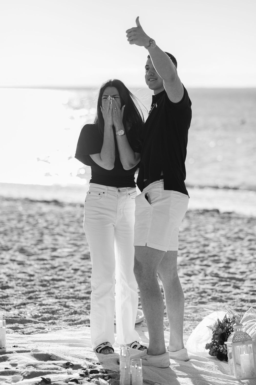 Black and white beach proposal photo with emotional woman covering mouth and man giving thumbs up.