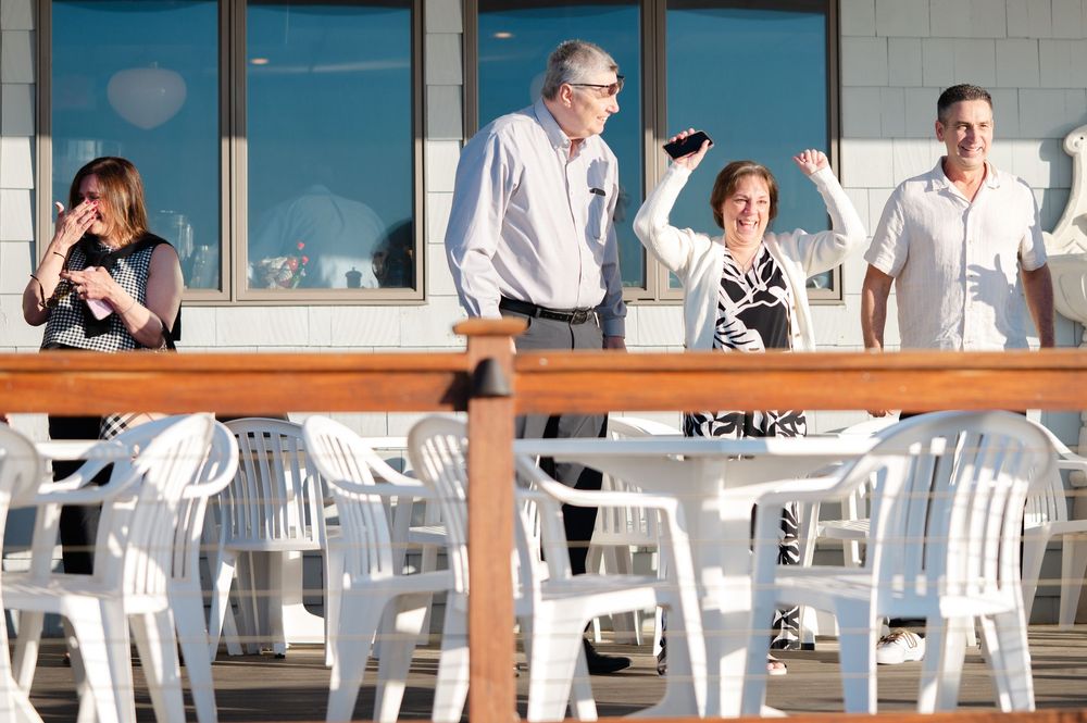 Four adults socializing on an outdoor patio with white plastic chairs and wooden railings on a sunny day.
