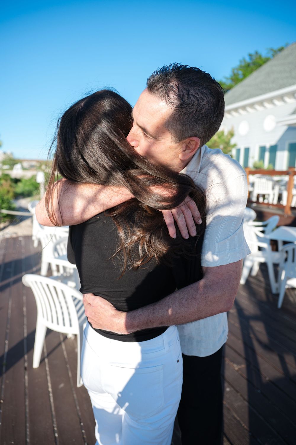 Couple sharing a tender hug on a sunny outdoor deck with white chairs and blue sky background.