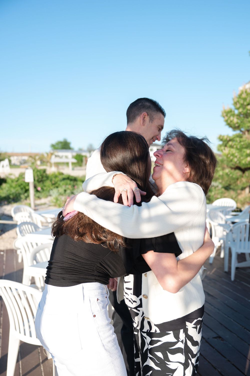 Joyful group hug on sunny outdoor deck with man in white jacket and two women celebrating.