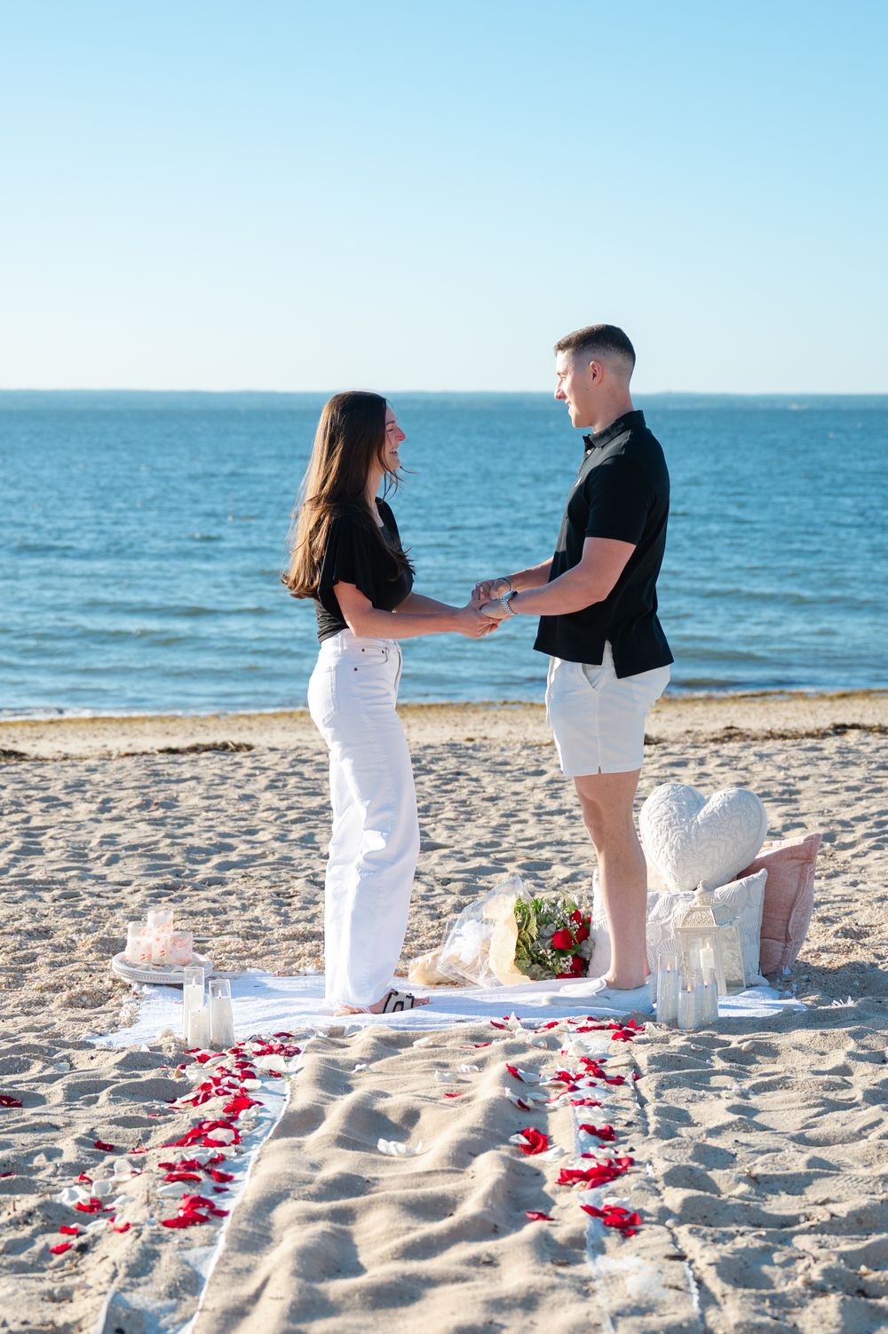 Couple holding hands on a romantic beach proposal setup with rose petals, lanterns, and ocean backdrop.