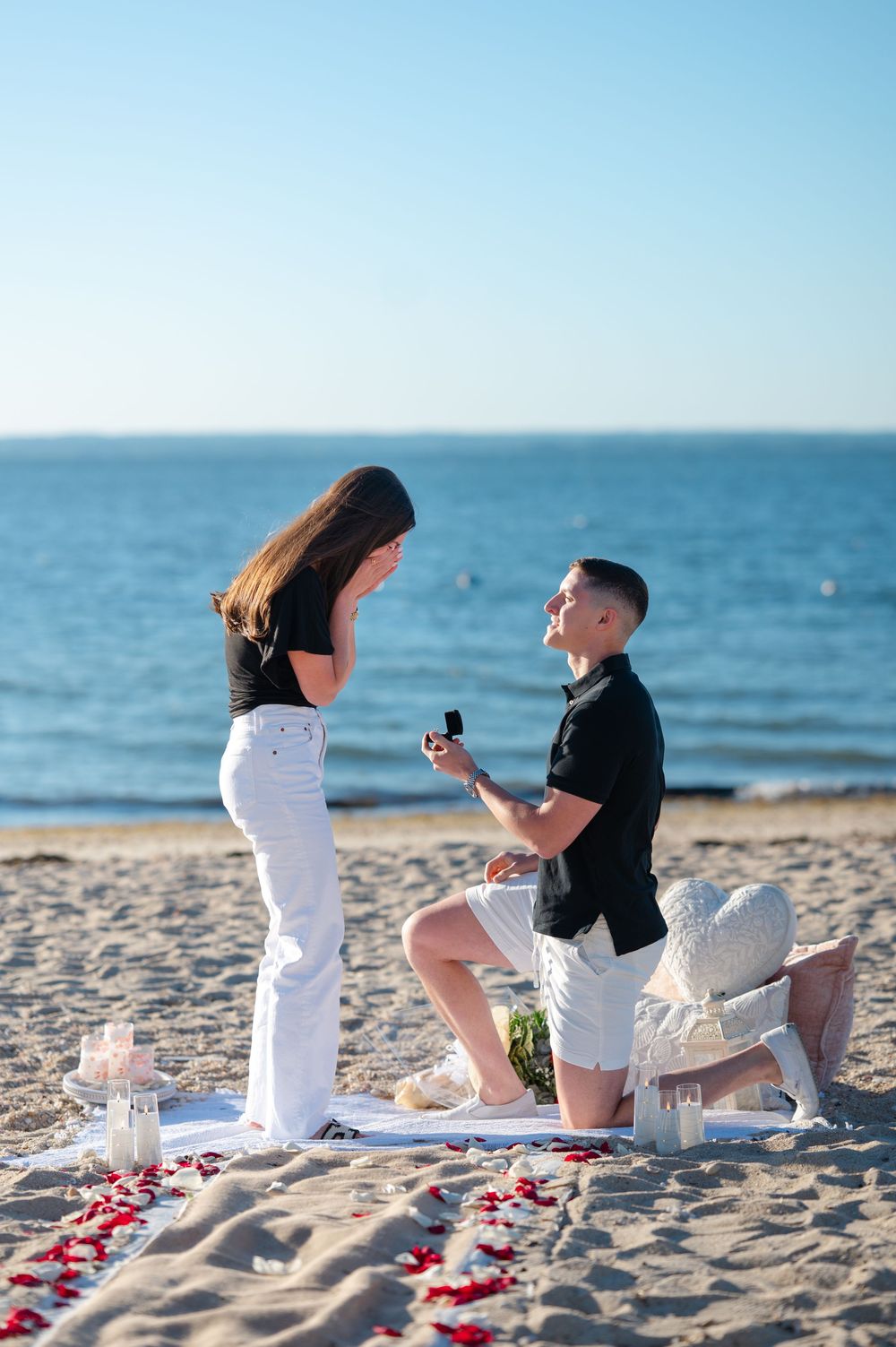 Man proposing on one knee to woman on sandy beach with rose petals, candles, and ocean backdrop on sunny day.