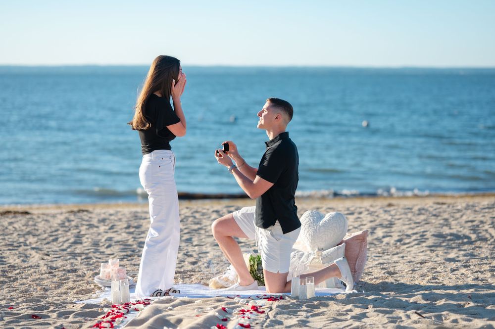 Man proposes to woman on sandy beach with rose petals, ocean backdrop, holding ring box on one knee.