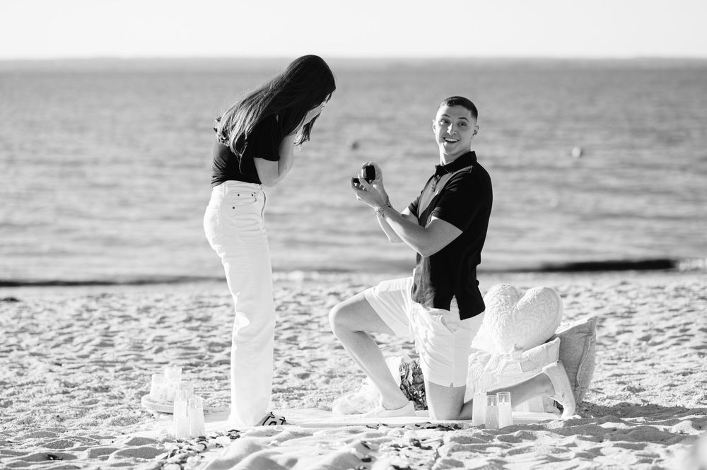 Man proposing on one knee to woman on beach with ring box, romantic black and white proposal photo.