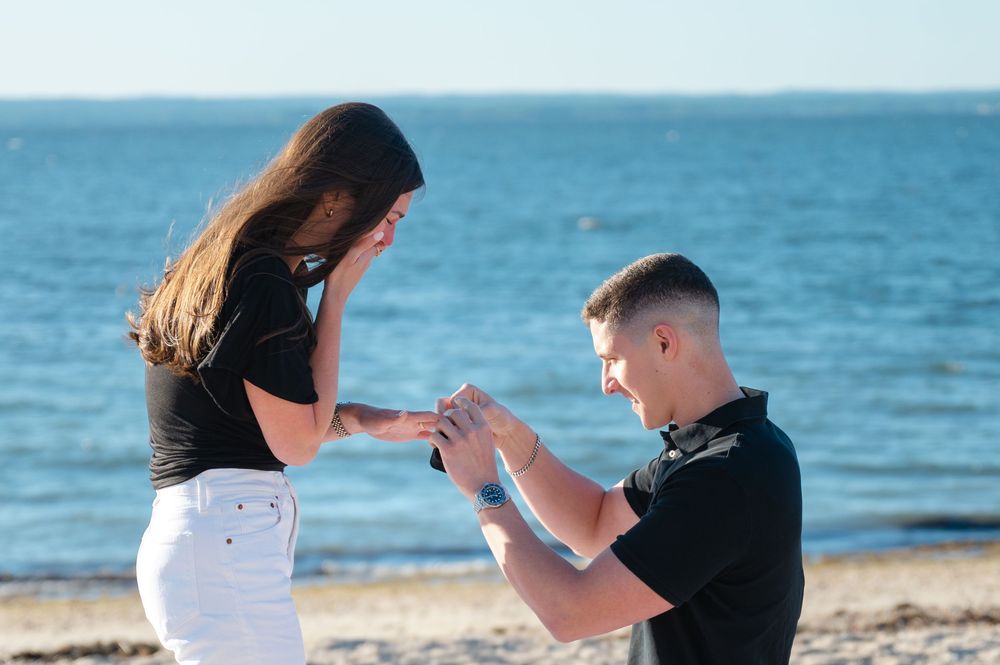 Man proposing to woman on a sandy beach by the ocean, placing a ring on her finger.