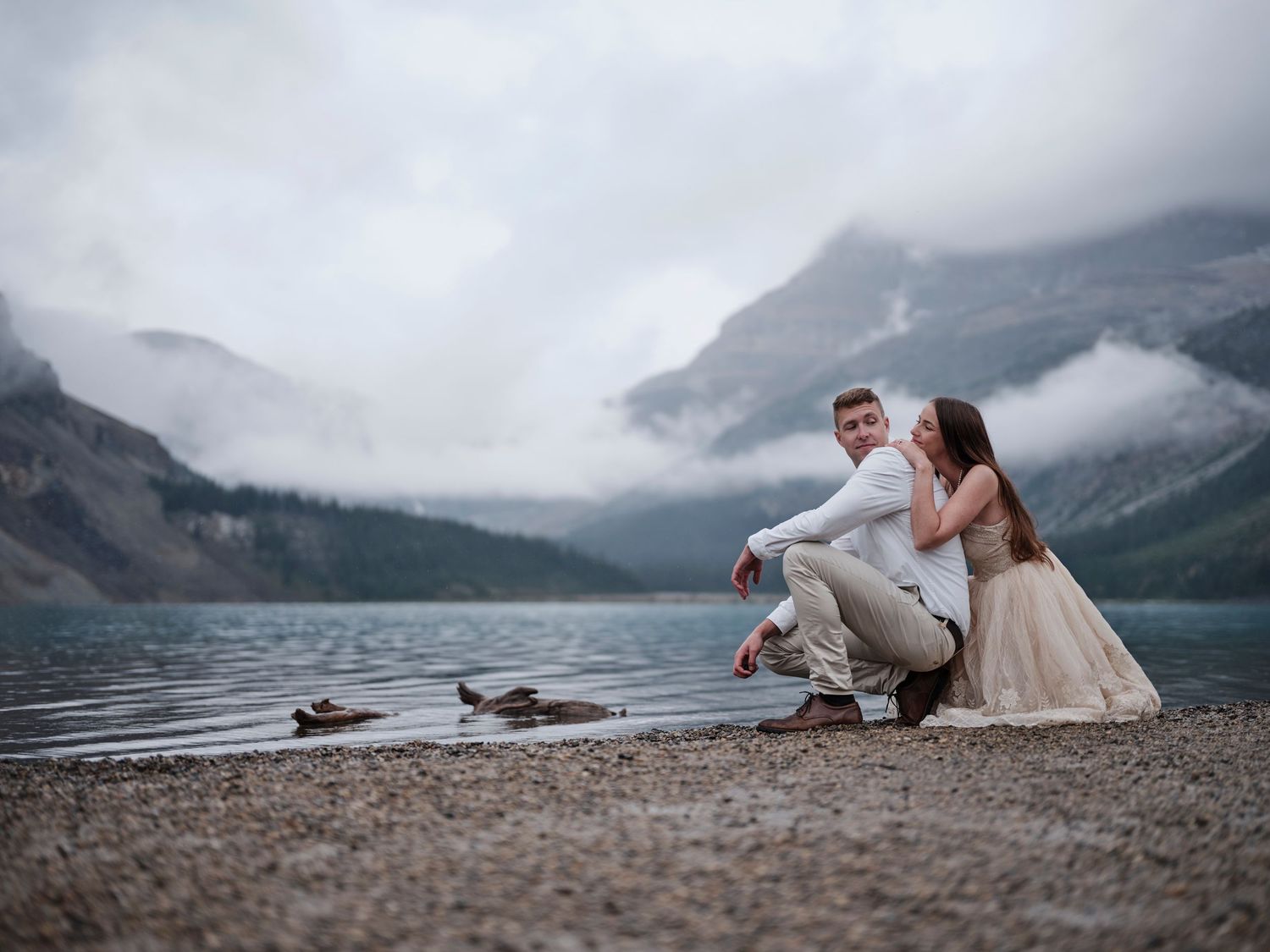 Couple sitting on rocky lakeshore with misty mountains and cloudy sky reflected in calm alpine lake.