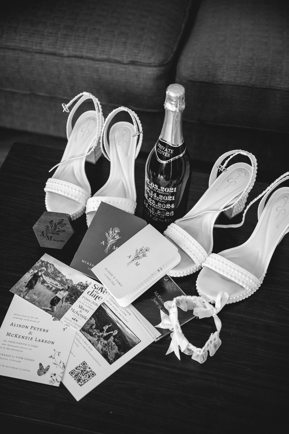 Black and white flat lay of bridal accessories including strappy heels, champagne bottle, wedding invitations, and a garter.