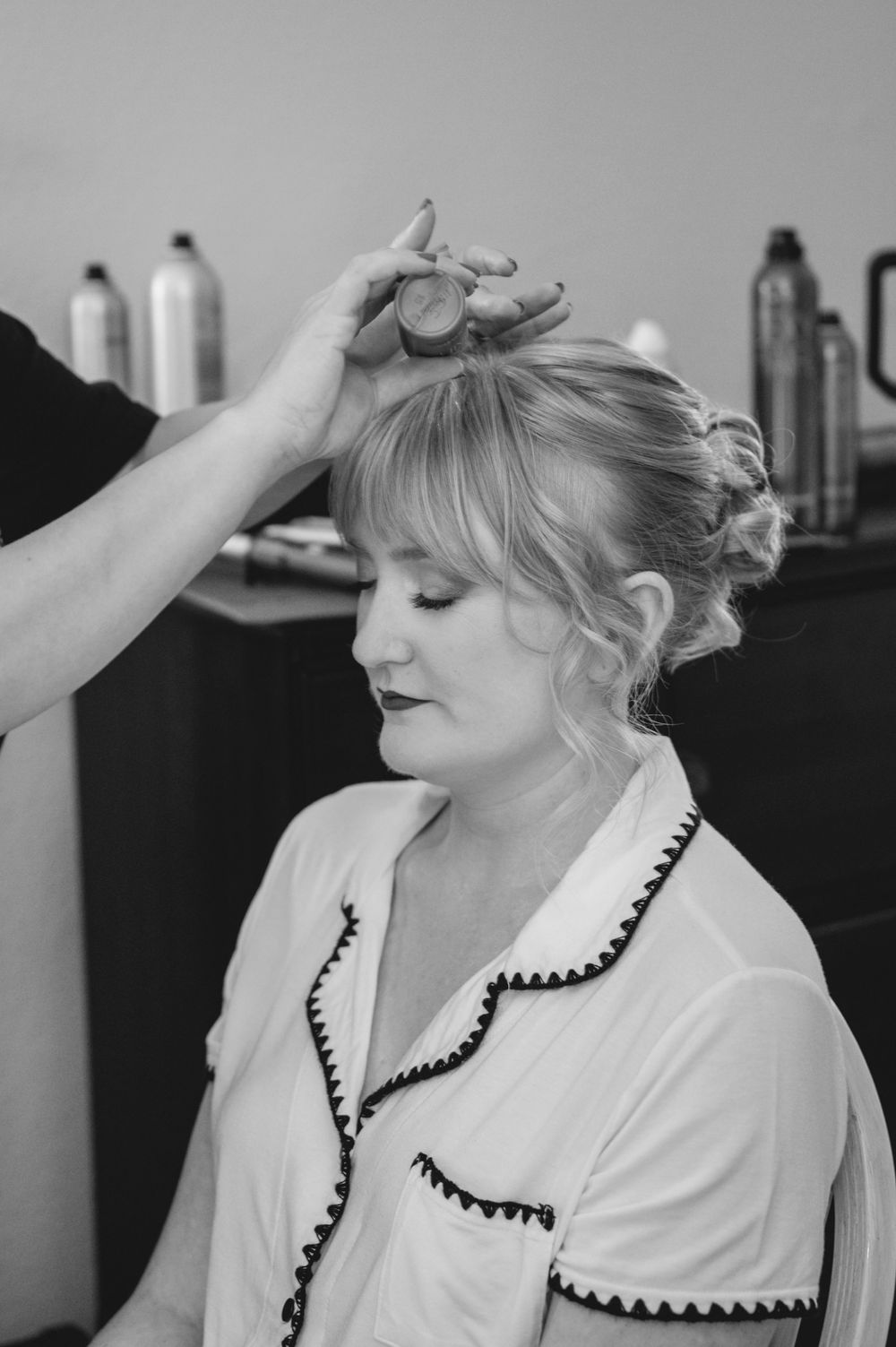Black and white photo of a bride having her updo hairstyle finished by a stylist in a bridal preparation room.
