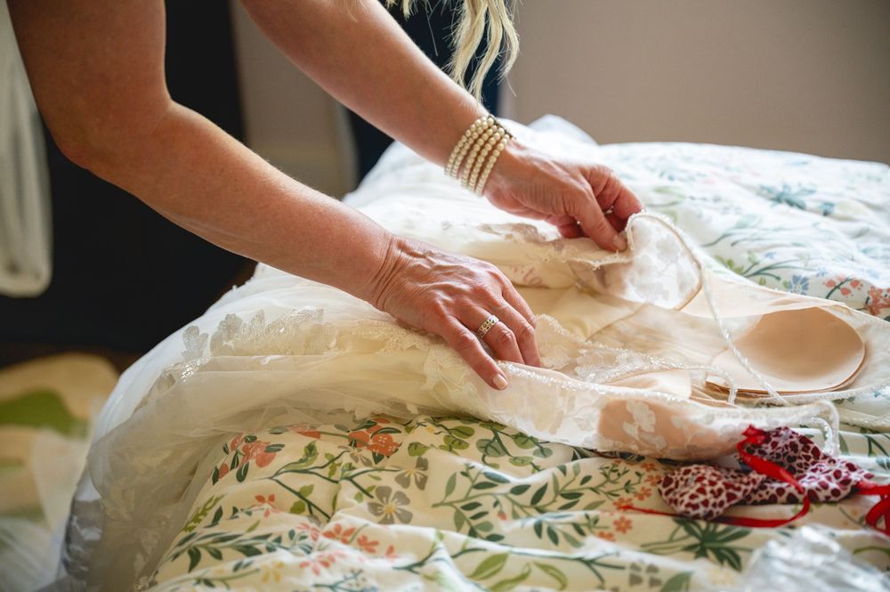 Woman with gold bracelets arranging a white lace wedding dress on a floral patterned bed.