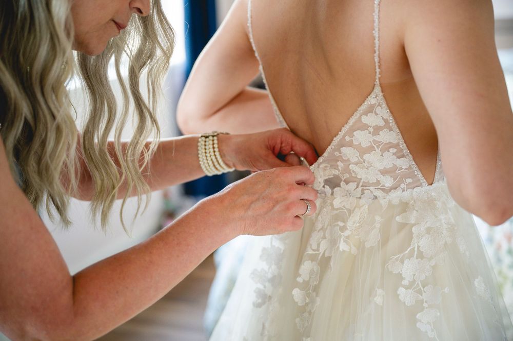 A bridesmaid fastening the back buttons of a bride's lace wedding gown before the ceremony.