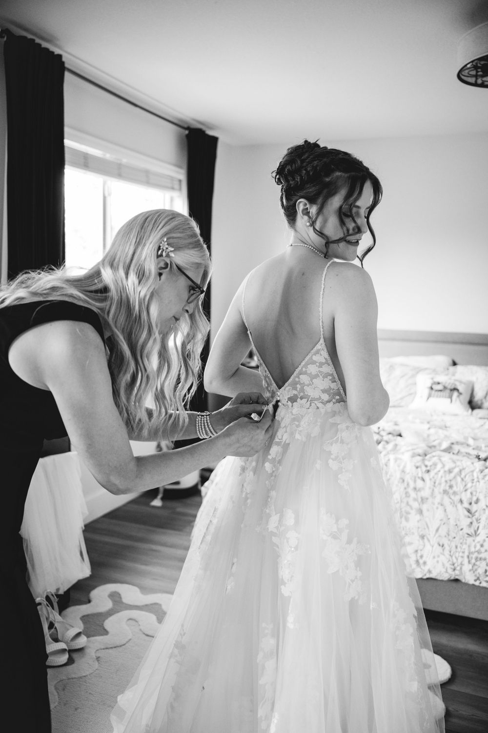 Black and white photo of a woman helping a bride fasten her elegant backless lace wedding gown in a bright room.