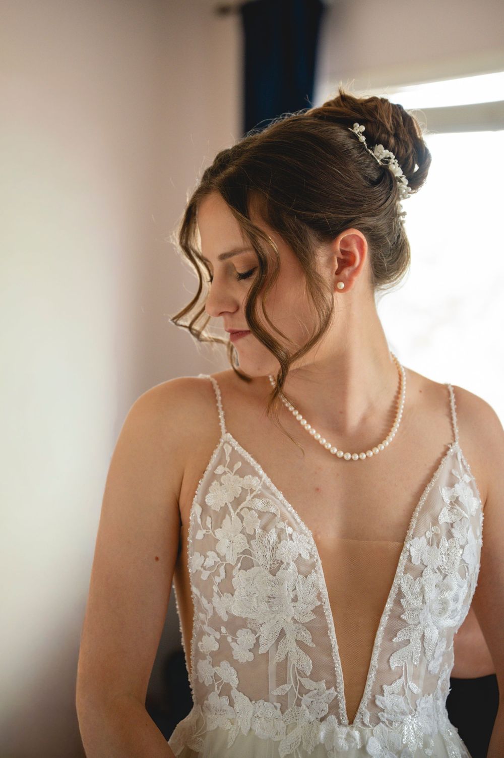 Bride in lace V-neck wedding gown with pearl necklace and floral hair accessory gazing down softly.