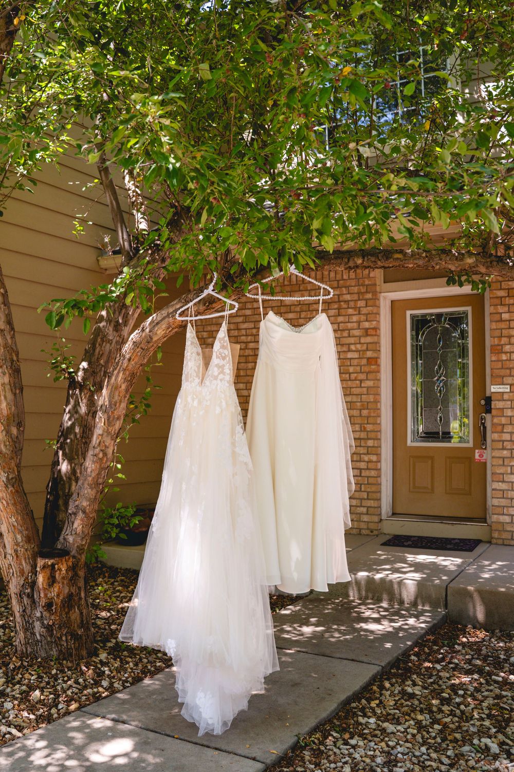 Two white wedding dresses hanging on hangers from a tree branch outside a brick home entrance.