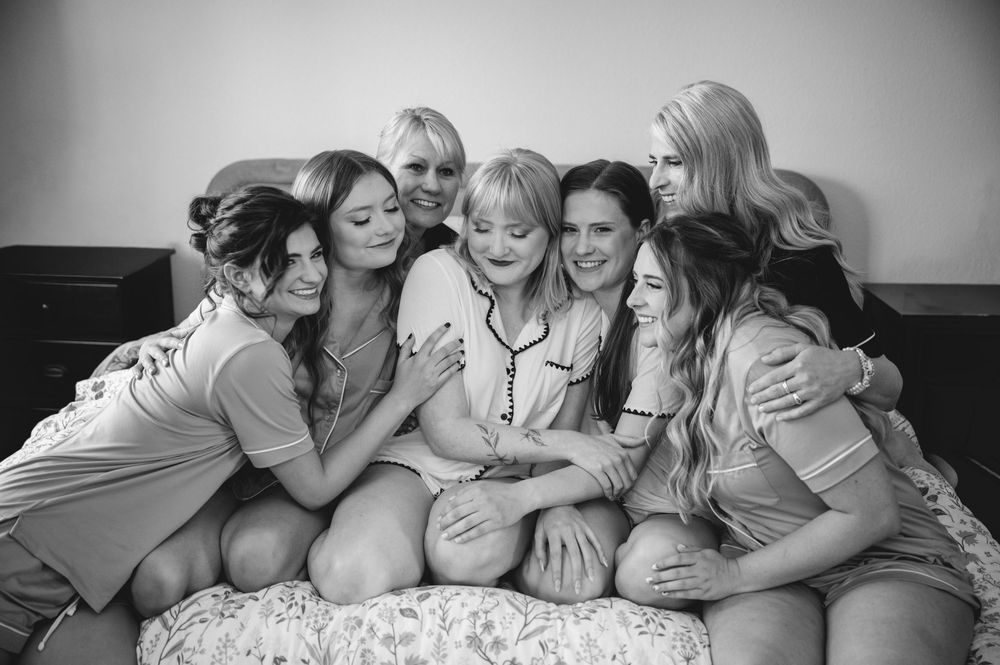 Black and white photo of seven women smiling and embracing together on a bed in a cozy, joyful group setting.