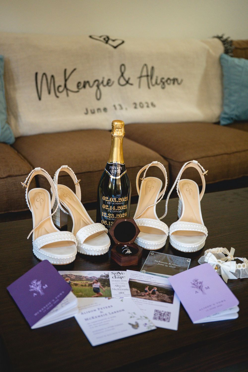 Wedding flat lay with white heeled sandals, champagne bottle, ring box, and purple programs for McKenzie and Alison, June 13, 2026.