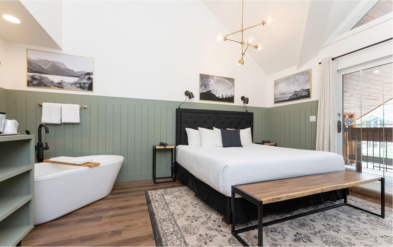 Modern hotel bedroom with soaking tub, green shiplap walls, king bed, hardwood floors, and mountain artwork.