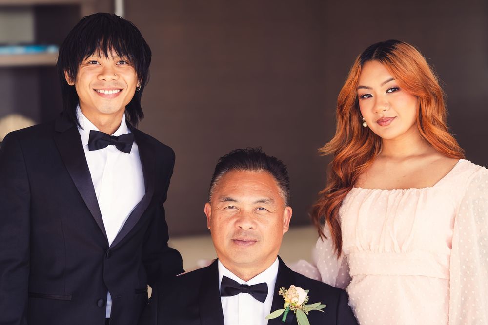 Three formally dressed individuals pose together at a wedding, two men in tuxedos and a woman in a pink dress.