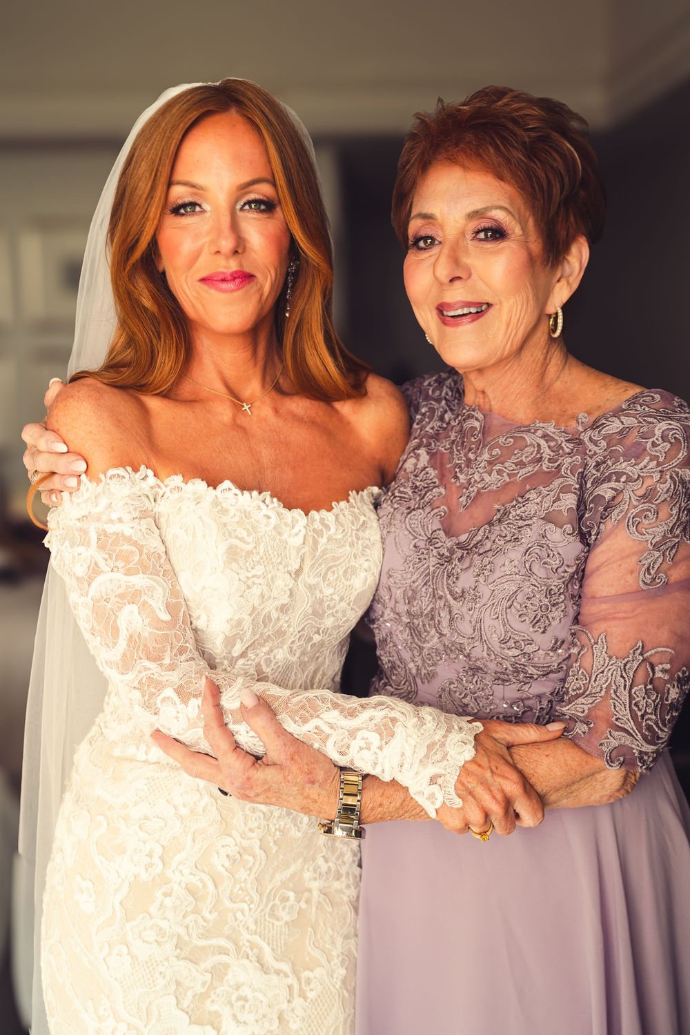 A bride in a white lace gown and veil poses with an older woman in a lavender dress at a wedding.