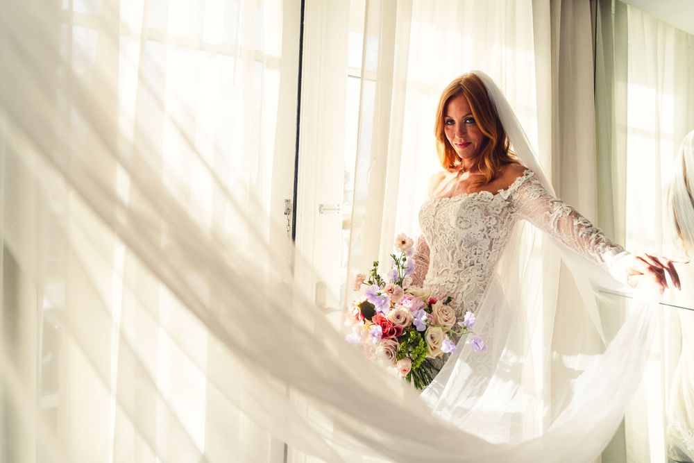 Bride in lace wedding gown holding colorful bouquet standing by sunlit window with flowing white curtains.