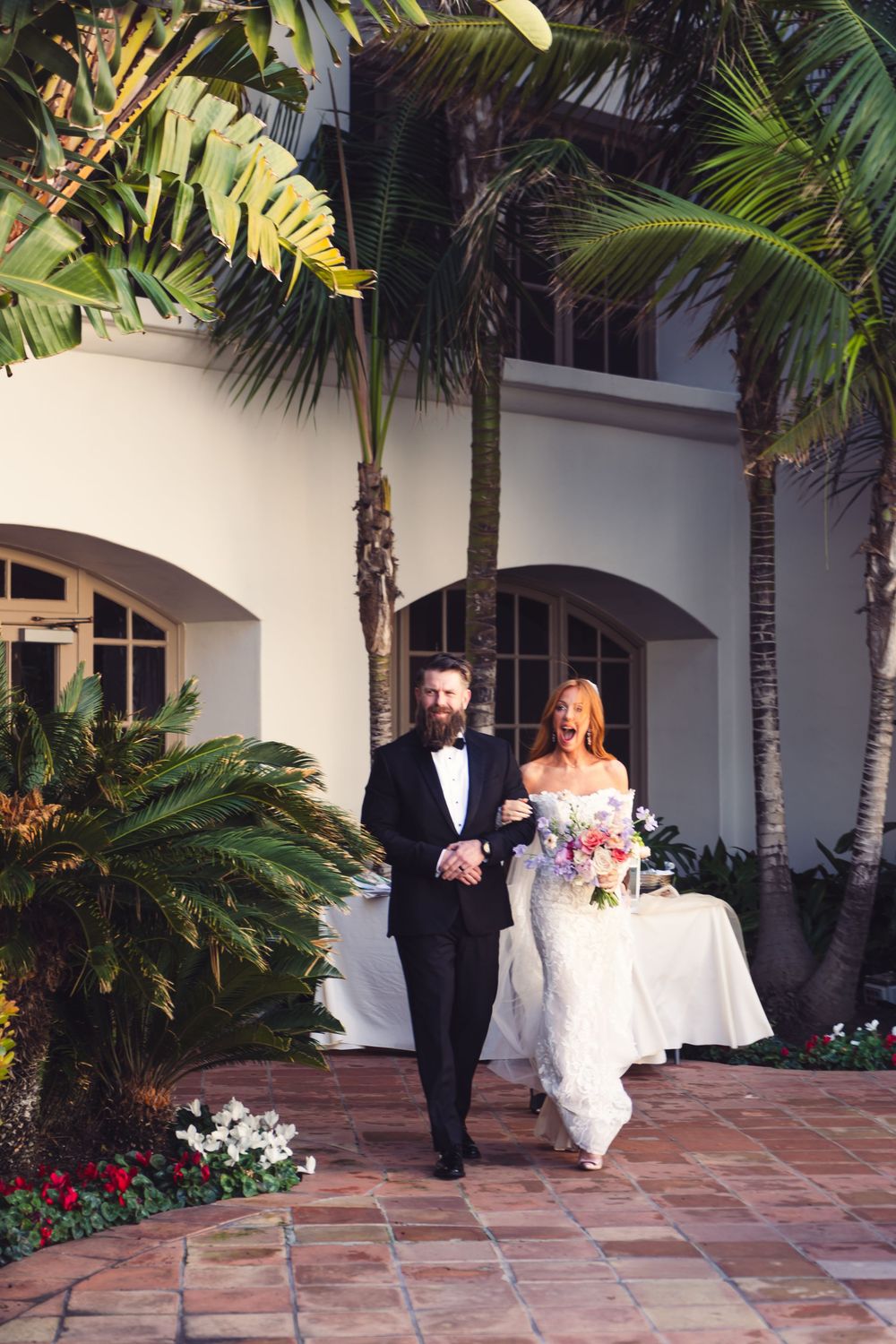 Bride and groom walk together outdoors past palm trees at a tropical wedding venue, bride holding colorful bouquet.