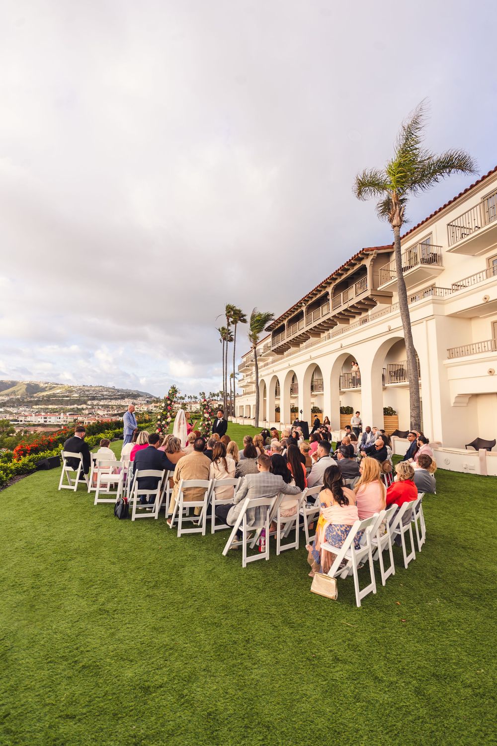 Outdoor wedding ceremony on green lawn beside elegant white Spanish-style venue with palm trees and ocean view.
