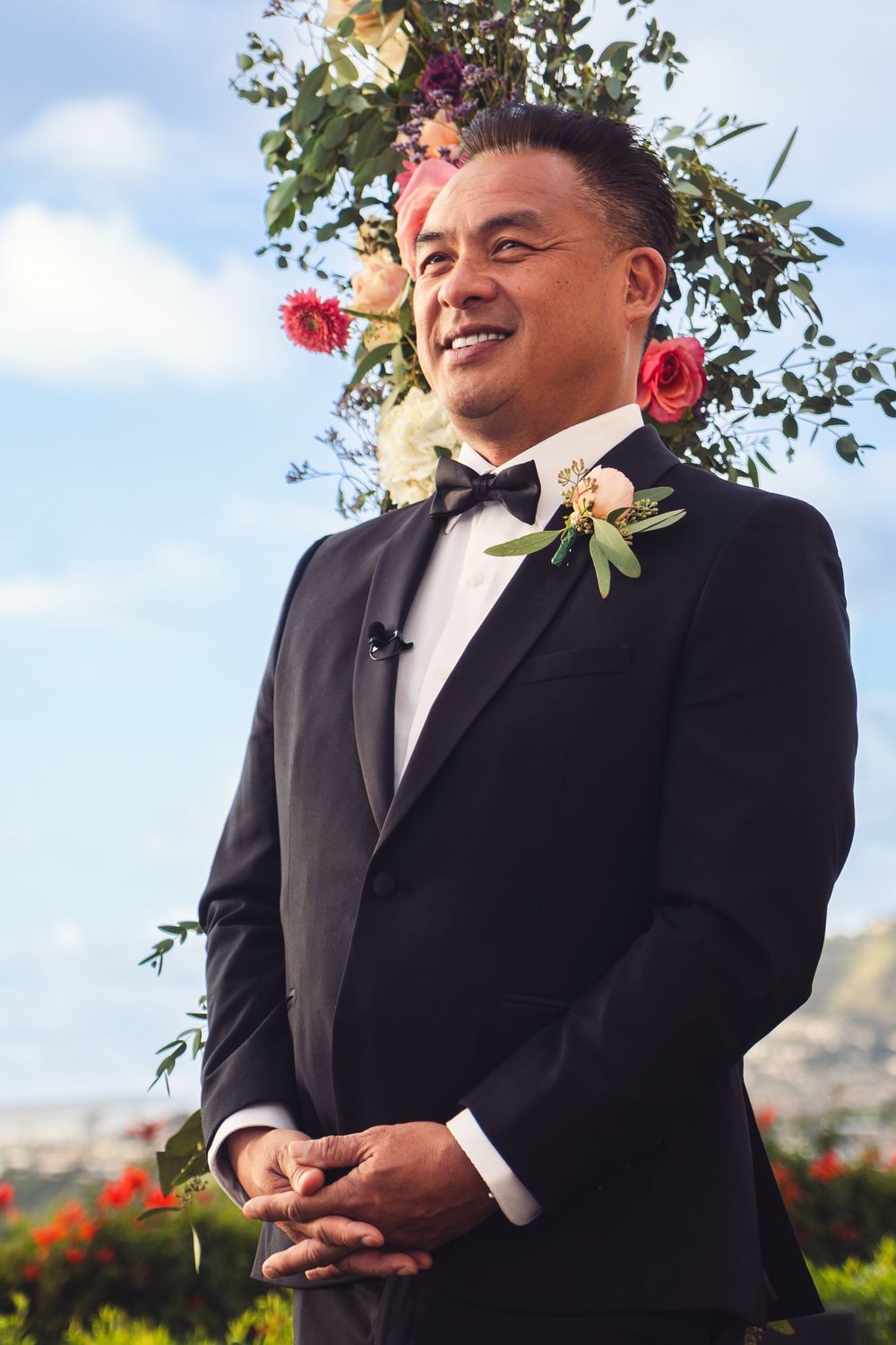 Smiling groom in black tuxedo stands at outdoor wedding altar adorned with colorful floral arrangement.