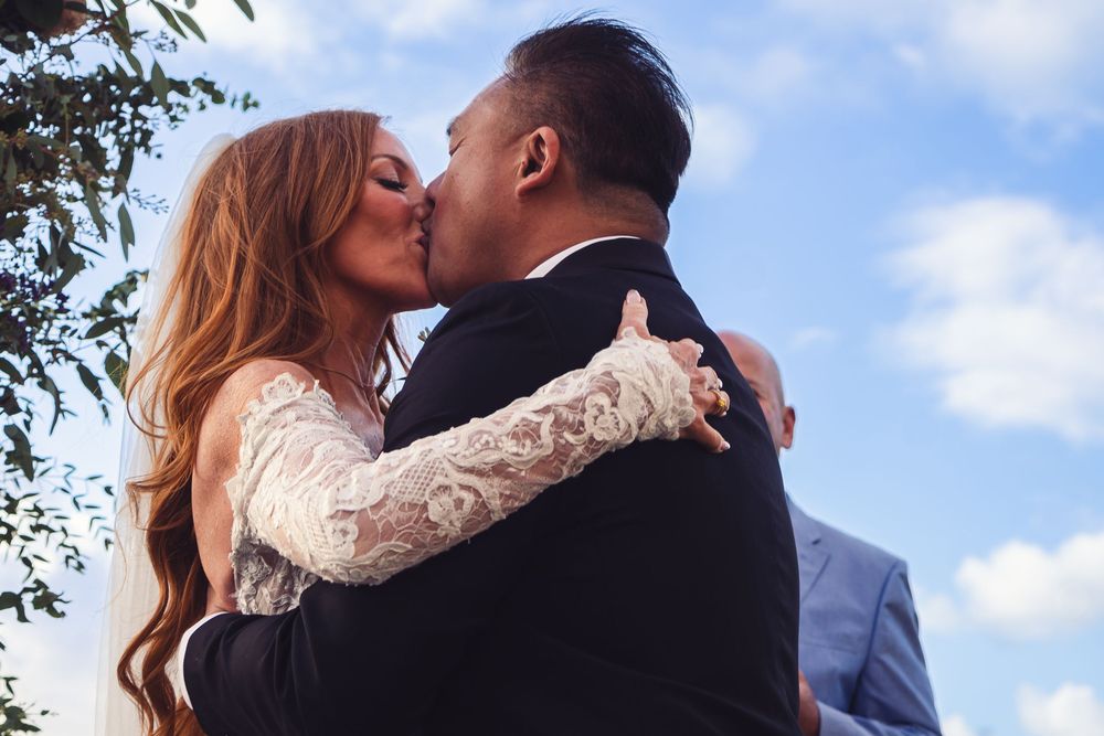Bride and groom share romantic first kiss at outdoor wedding ceremony under blue sky.