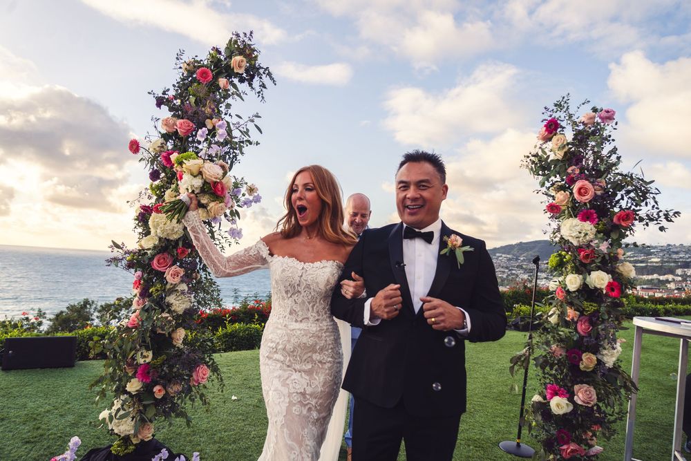 Joyful bride and groom celebrating at outdoor wedding ceremony with floral arch and ocean view backdrop.