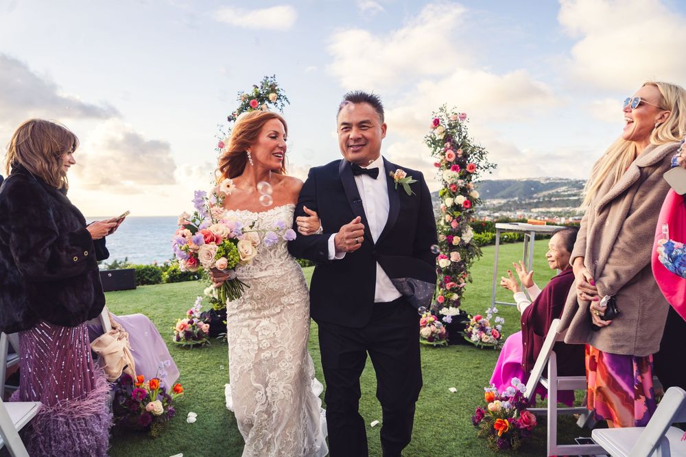 Bride and groom walk down the aisle at an outdoor wedding ceremony with colorful floral arch and ocean views.