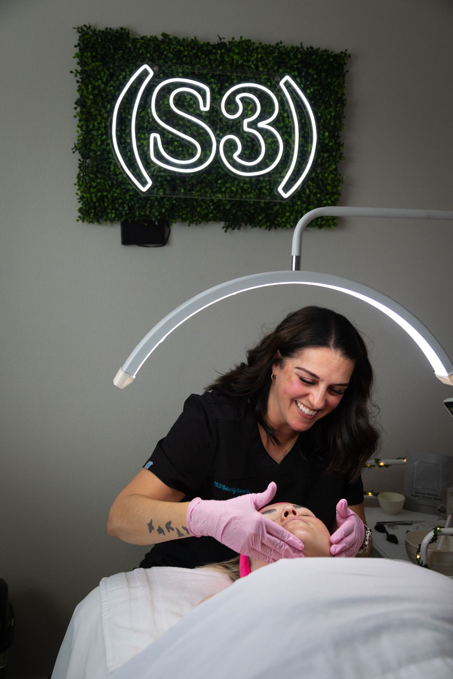 Esthetician in black uniform and pink gloves performing a facial treatment under an arch lamp at S3 beauty salon.
