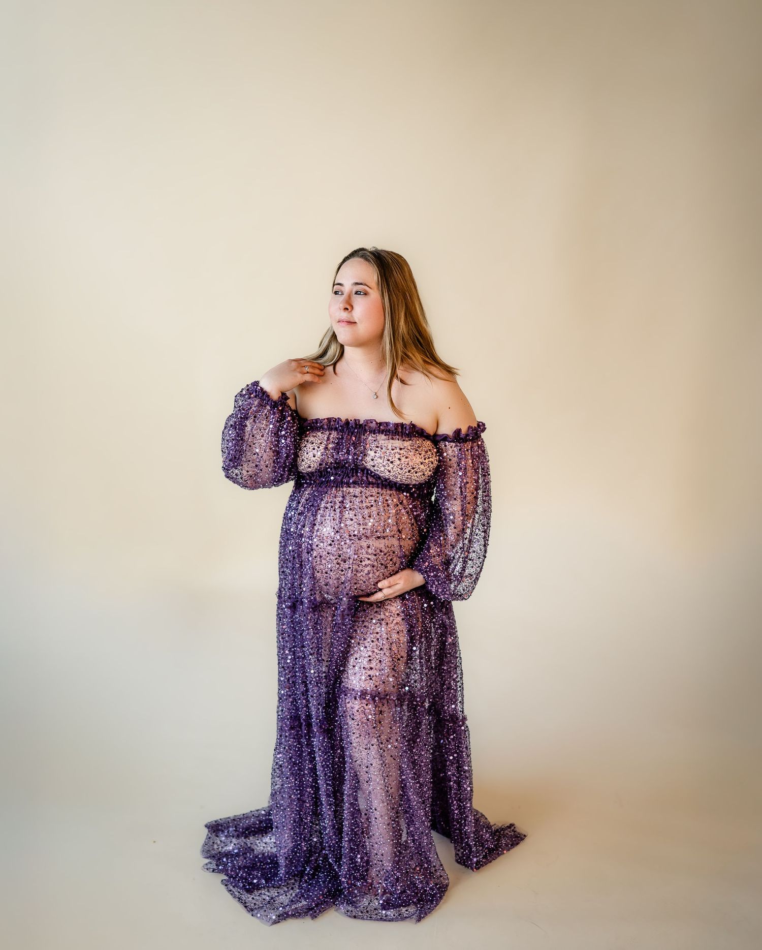 Pregnant woman in sheer purple off-shoulder maternity gown posing gracefully in a professional studio photo.