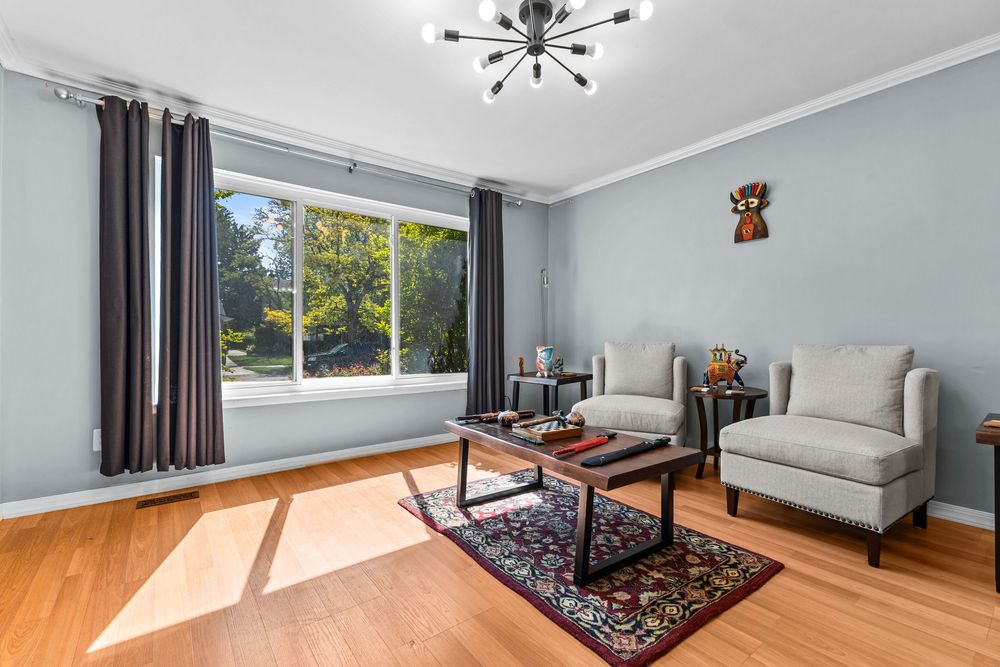 Sunlit living room with hardwood floors, gray walls, crown molding, large window, and modern chandelier.